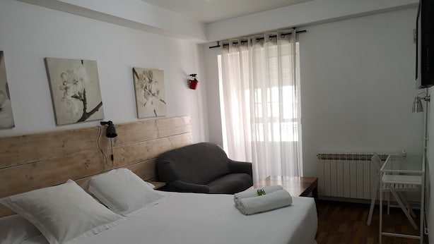 A neatly made bed with white linens, a grey armchair, a small table with a bottle and a towel, and a radiator. - Hostal Carbonara (A Coruña)