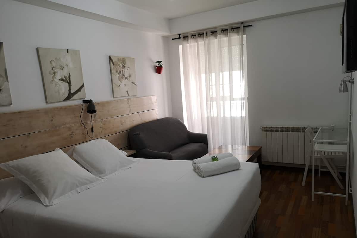 Una cama bien hecha con sábanas blancas, un sillón gris, una mesita con una botella y una toalla, y un radiador.