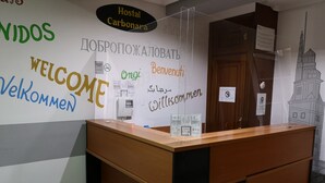 A reception desk with a wall displaying multilingual welcome signs and a poster of a building. - Hostal Carbonara (A Coruña)