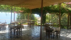 Un patio al aire libre con mesas y sillas, cubierto por una estructura de madera y rodeado de follaje.