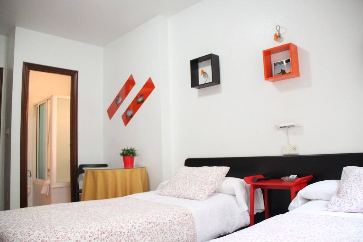 Quarto de hotel com duas camas, uma mesa pequena e um vaso de planta.
