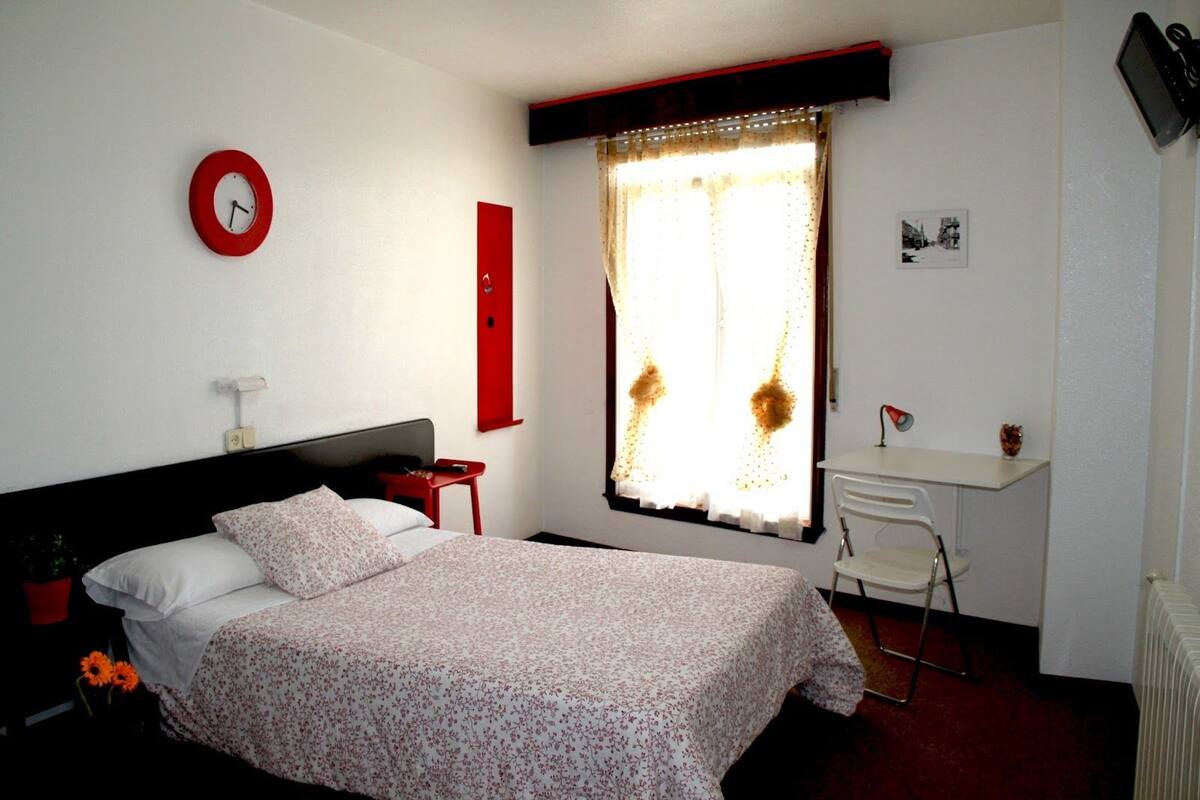 Quarto com cama, escrivaninha, cadeira, relógio e janela com cortinas.