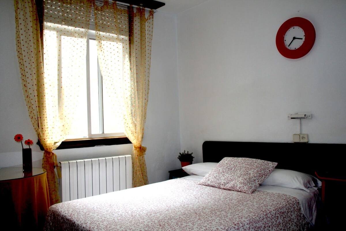 Um quarto com uma cama, uma janela com cortinas, um relógio na parede e uma mesinha de cabeceira com uma planta.