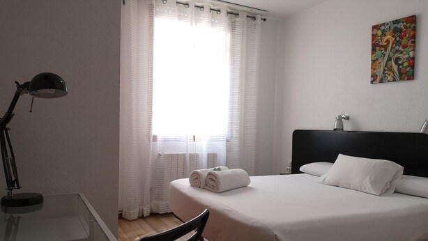 A hotel room with a bed, a desk, a chair, a lamp, and a window with curtains. - Pension Las Rias (A Coruña)