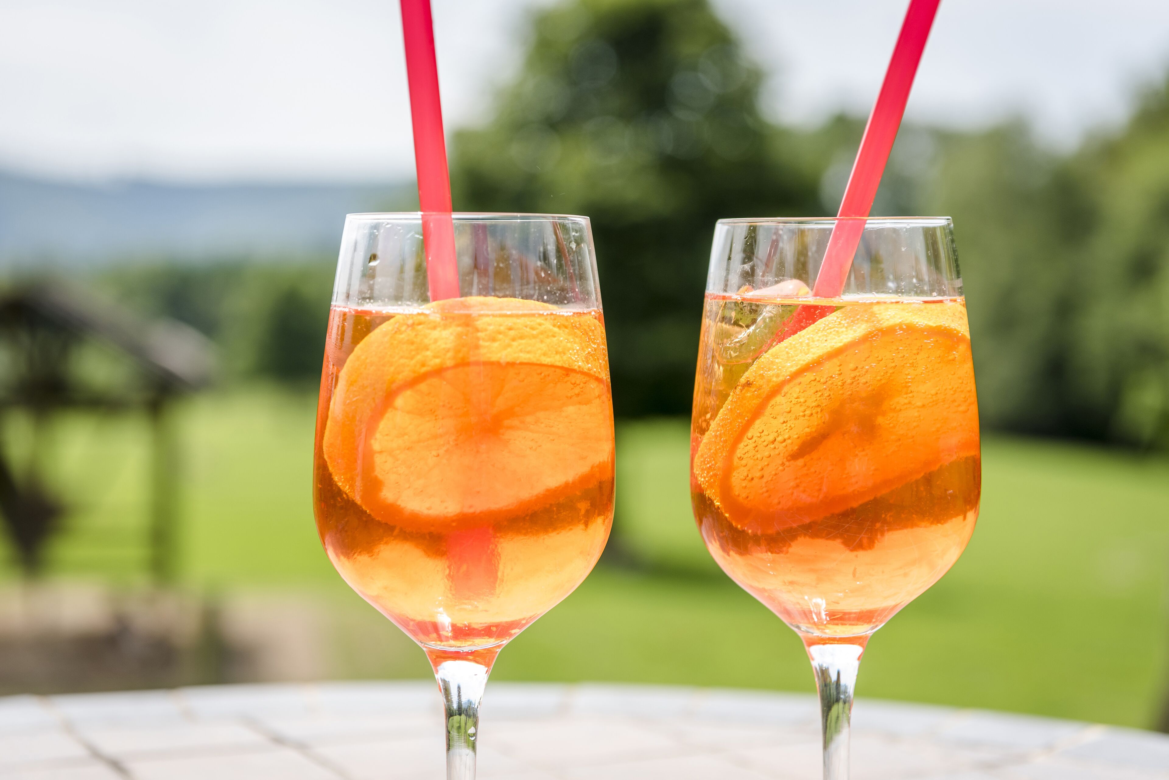 Dos vasos de una bebida transparente con rodajas de naranja y una pajita rosa, sobre una mesa con un fondo exterior borroso.