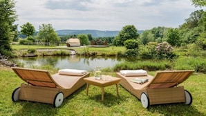 Deux fauteuils de jardin en bois, équipés de roulettes, sont placés face à un étang. Une petite table avec un verre d’eau se trouve à proximité, et on aperçoit une maison et des arbres au loin.