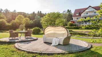 A round wooden deck with a beige sofa and two chairs, surrounded by a garden with a house in the background.