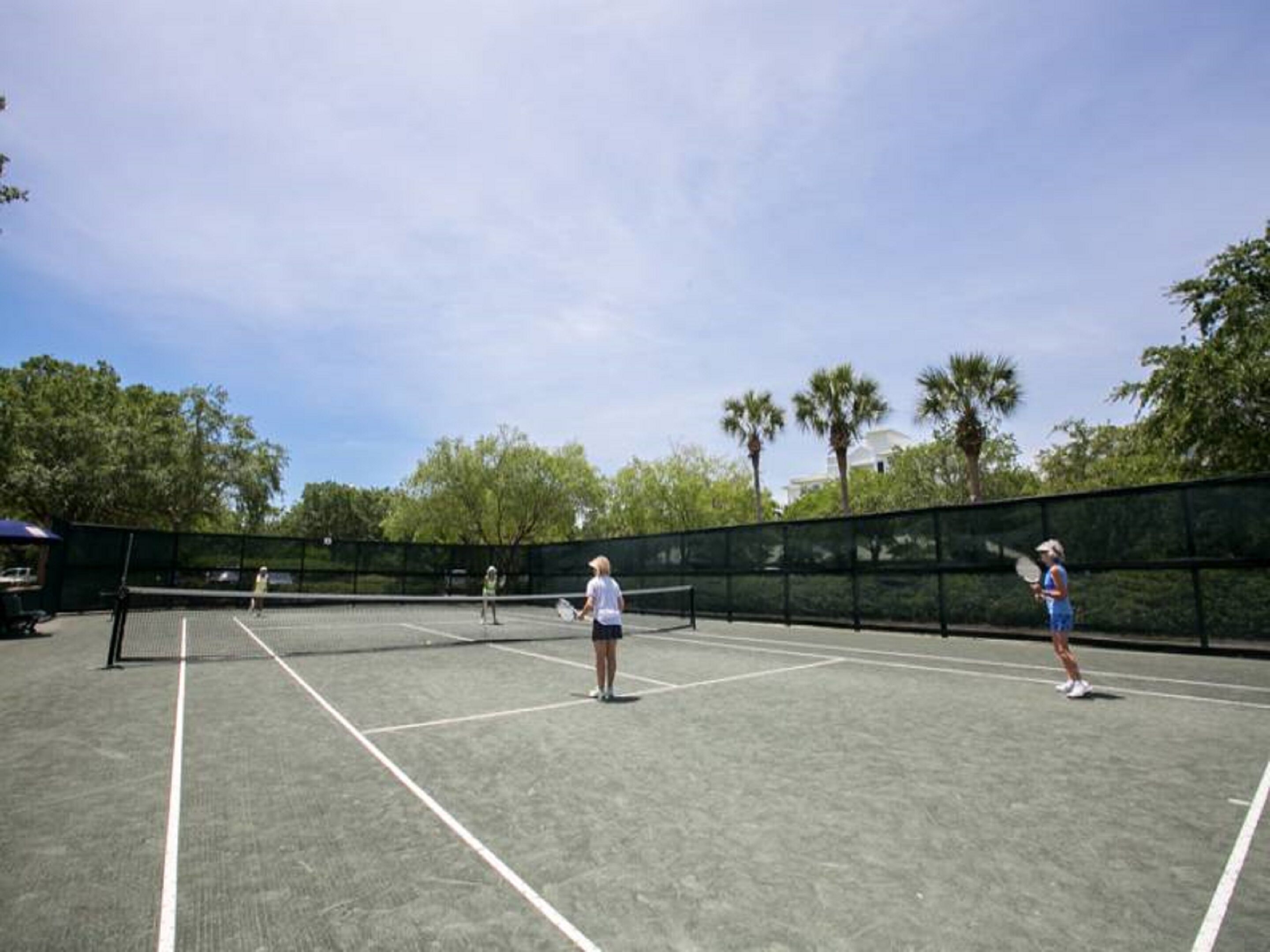 An outdoor tennis court with a net, surrounded by trees and a clear sky.