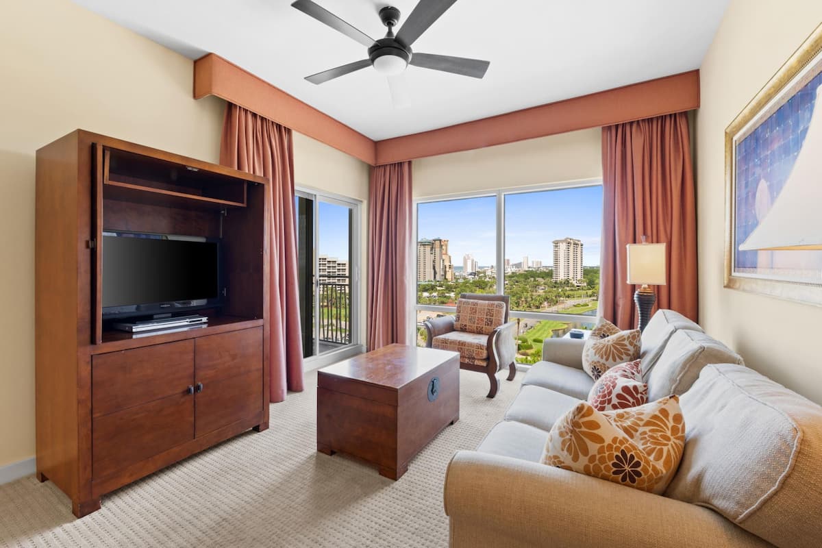 Quarto de hotel com uma TV de tela plana grande, um armário de madeira, um sofá com almofadas estampadas, uma mesa de centro e vista para a paisagem urbana através de grandes janelas.
