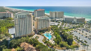 A resort with multiple high-rise buildings, a central swimming pool, and a view of the ocean.