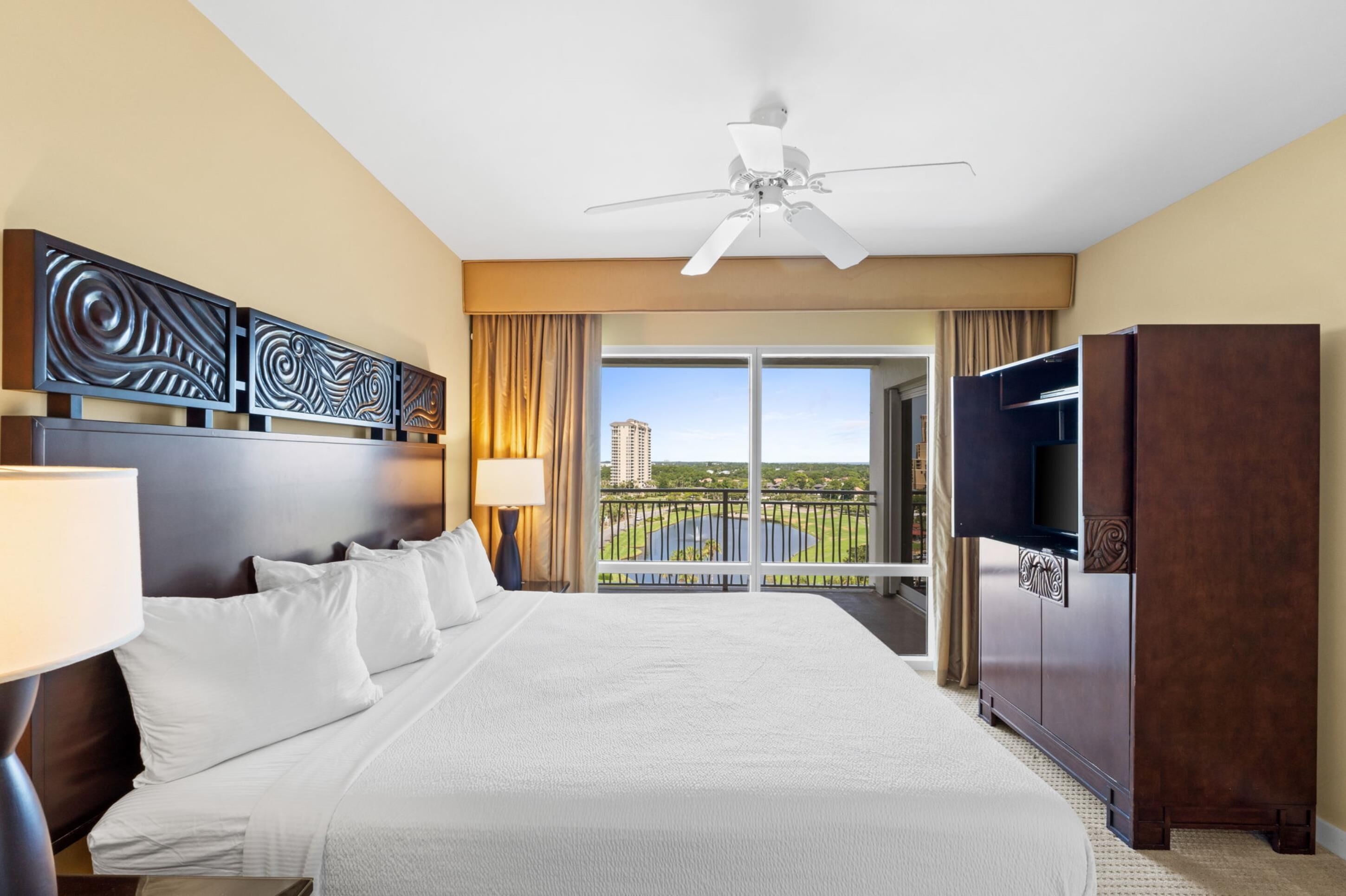 A hotel room with a large bed, a ceiling fan, a TV, and a balcony with a view of a cityscape.