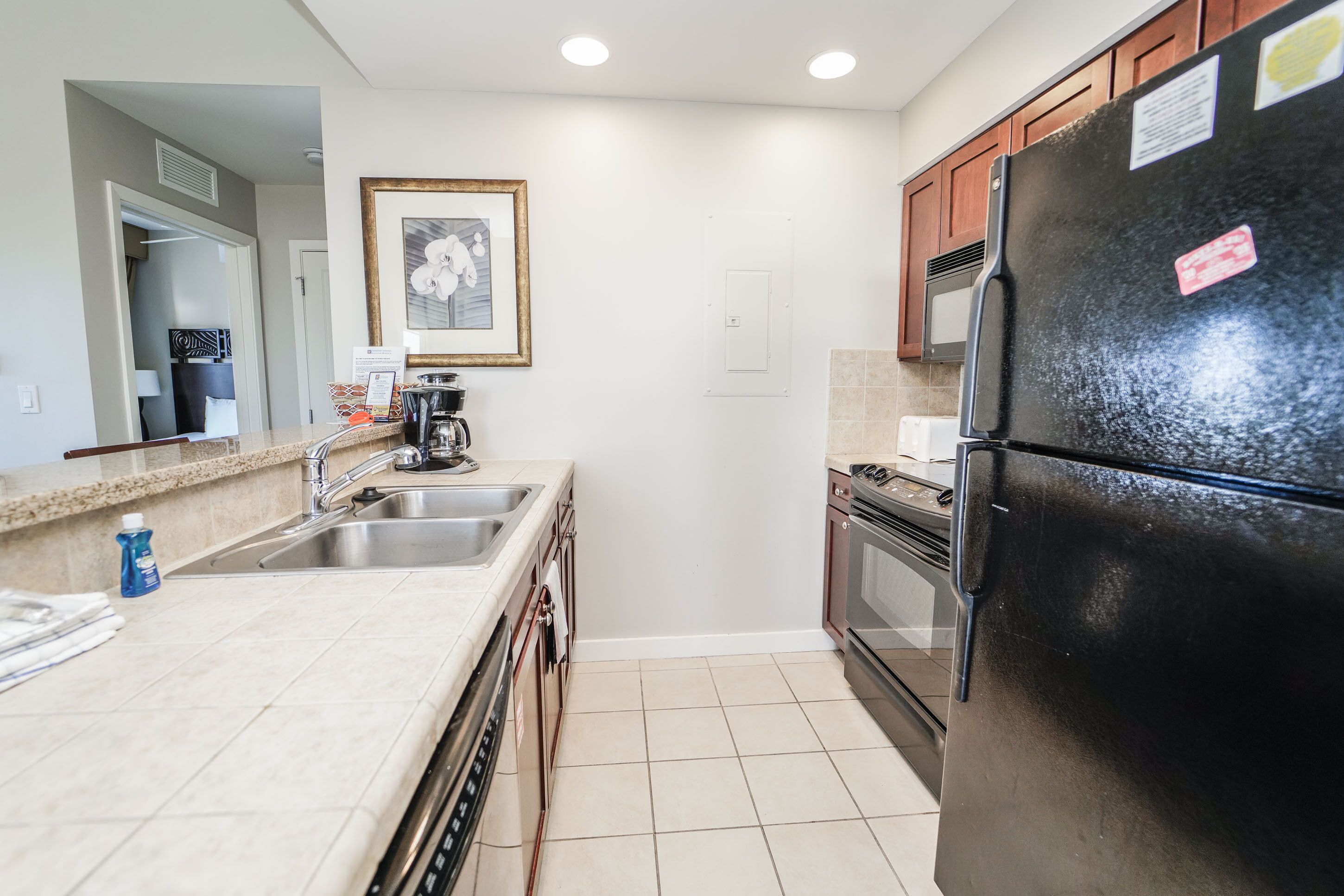 A kitchen with a black refrigerator, stainless steel sink, and a microwave. There is a framed picture on the wall and a door with a light switch.