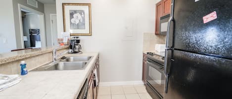A kitchen with a black refrigerator, stainless steel sink, and a microwave. There is a framed picture on the wall and a door with a light switch.
