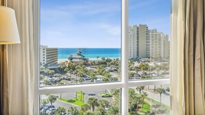 A hotel room with a large window overlooking a beach, parking area, and buildings.