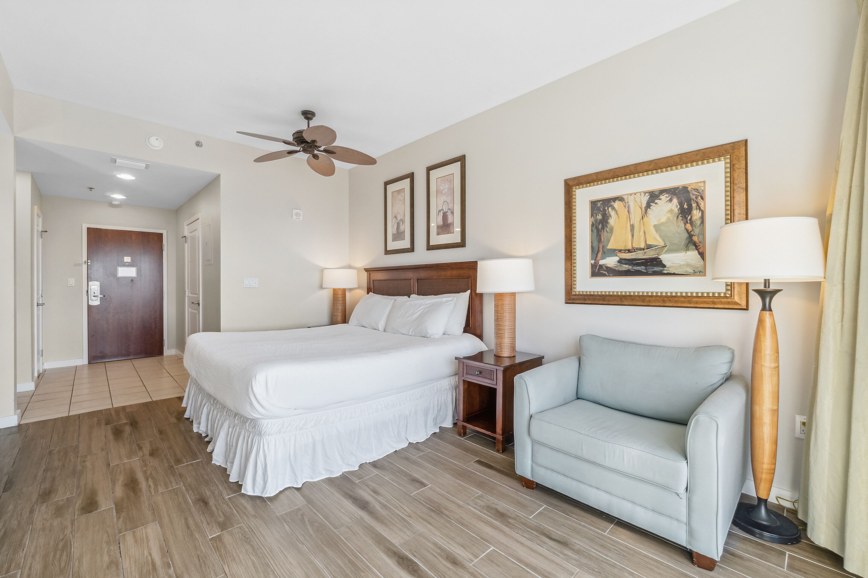 Une chambre d’hôtel avec un grand lit, un ventilateur de plafond, deux tableaux encadrés, un canapé et une lampe.