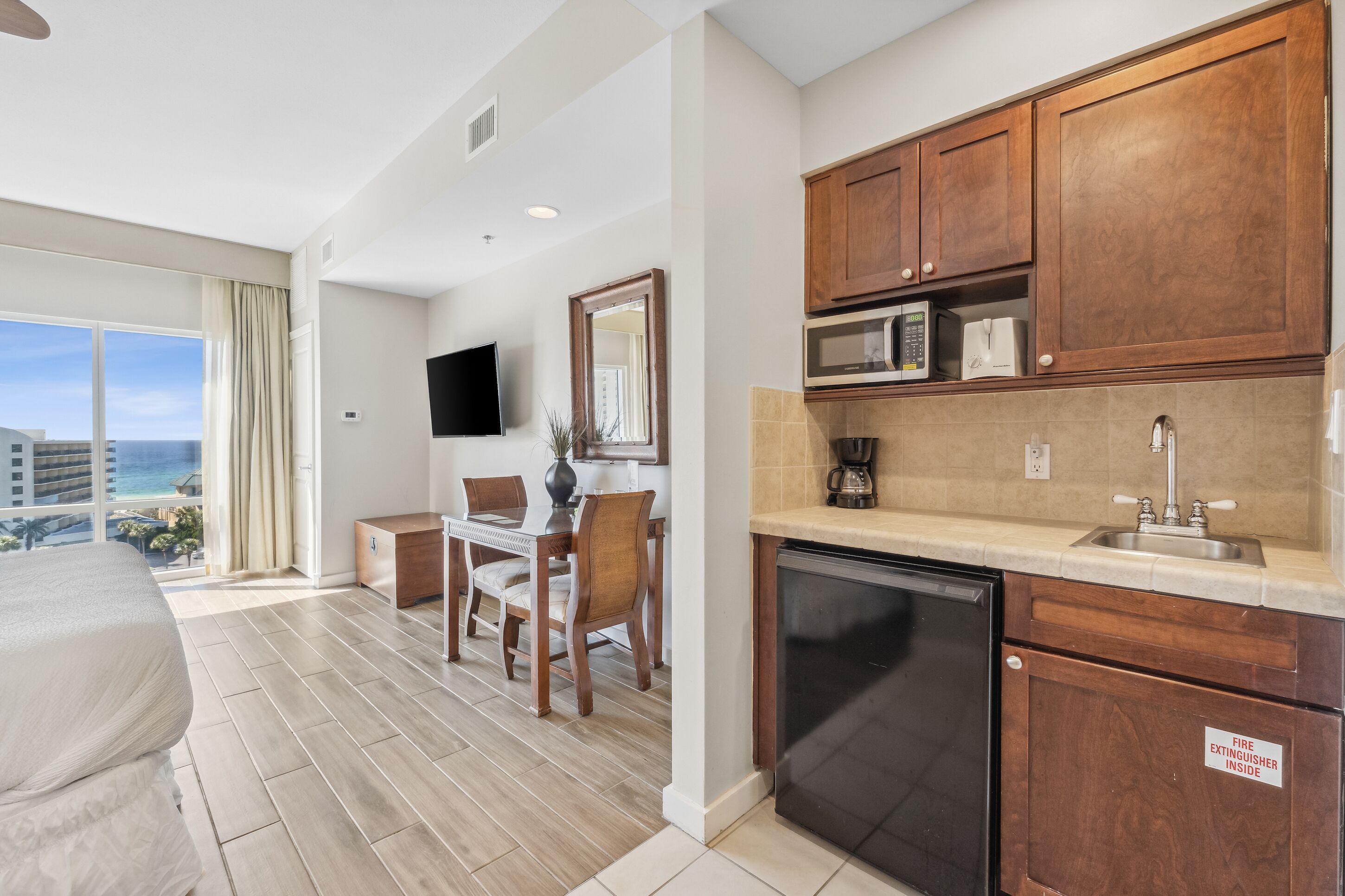 A hotel room with a bed, a dining area with a table and chairs, a kitchen with wooden cabinets and a microwave, and a view of the ocean.