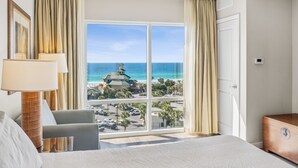 A hotel room with a large window offering a view of the beach and ocean, a bed, a chair, and a wooden dresser.