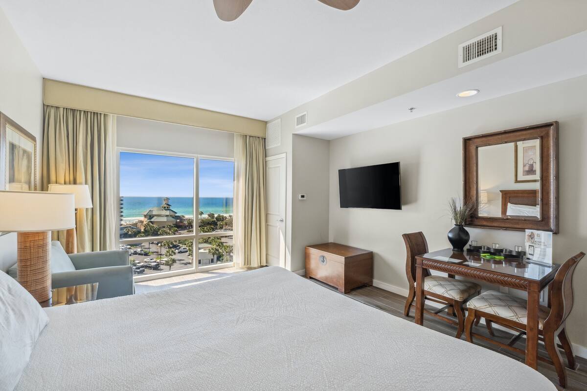 Quarto de hotel com uma cama grande, uma mesa com cadeira, televisão e vista para o oceano.