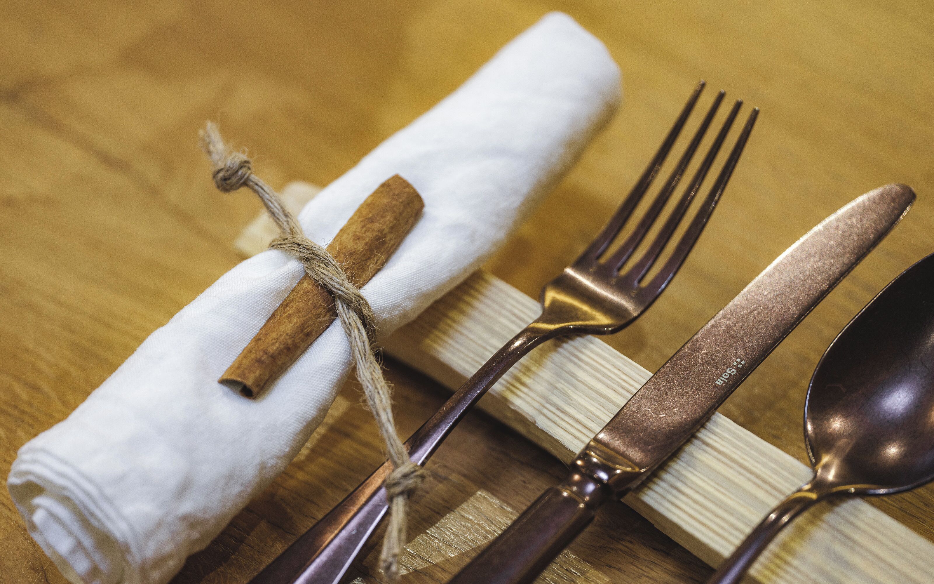 A neatly arranged table setting with a folded napkin tied with a cinnamon stick, accompanied by a fork, knife, and spoon.