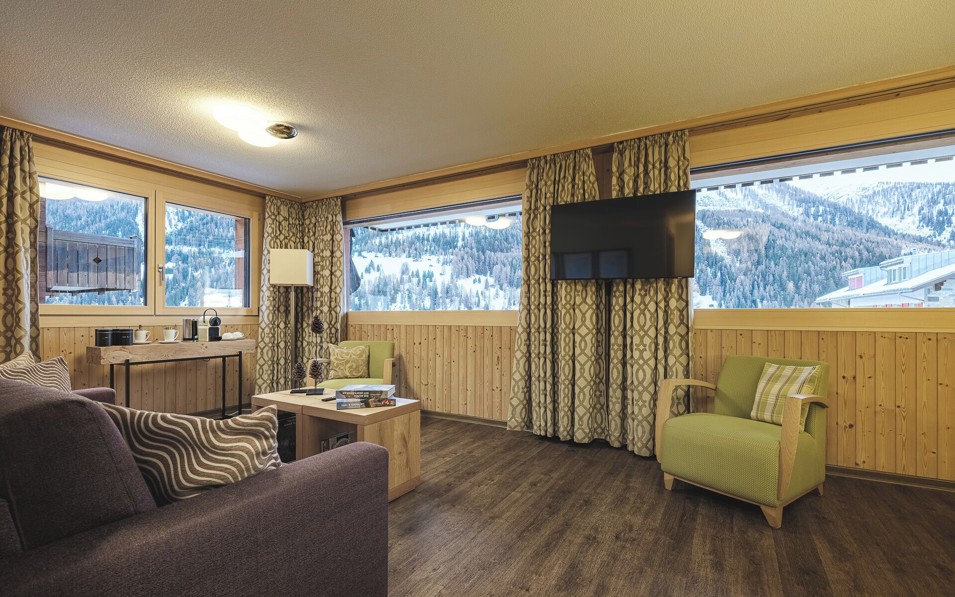 Un cuarto acogedor con piso de madera, un sofá, una silla, una mesita con una lámpara y un televisor. Amplias ventanas ofrecen vistas de montañas nevadas.