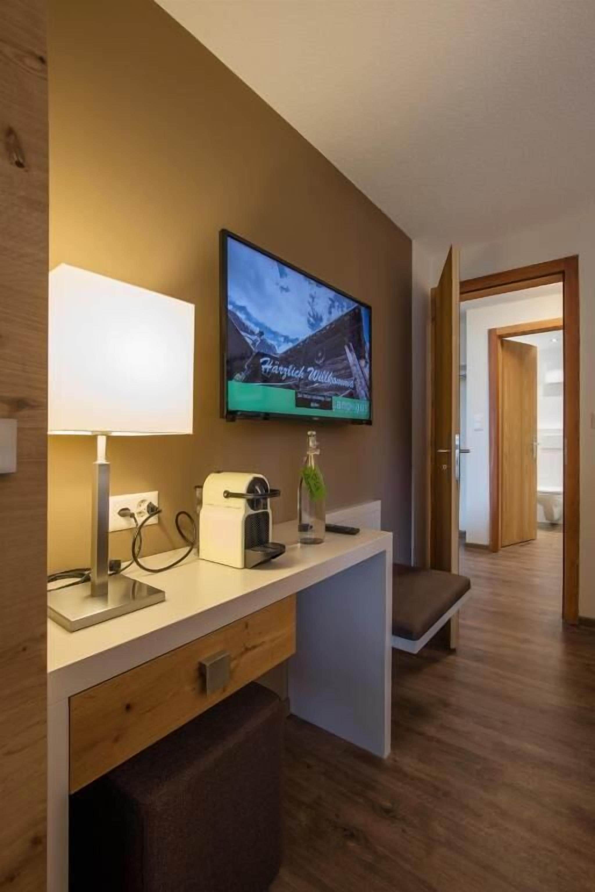 A hotel room with a television mounted on the wall, a bedside table with a lamp, a bottle, and a coffee maker.