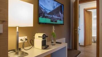 A hotel room with a television mounted on the wall, a bedside table with a lamp, a bottle, and a coffee maker.