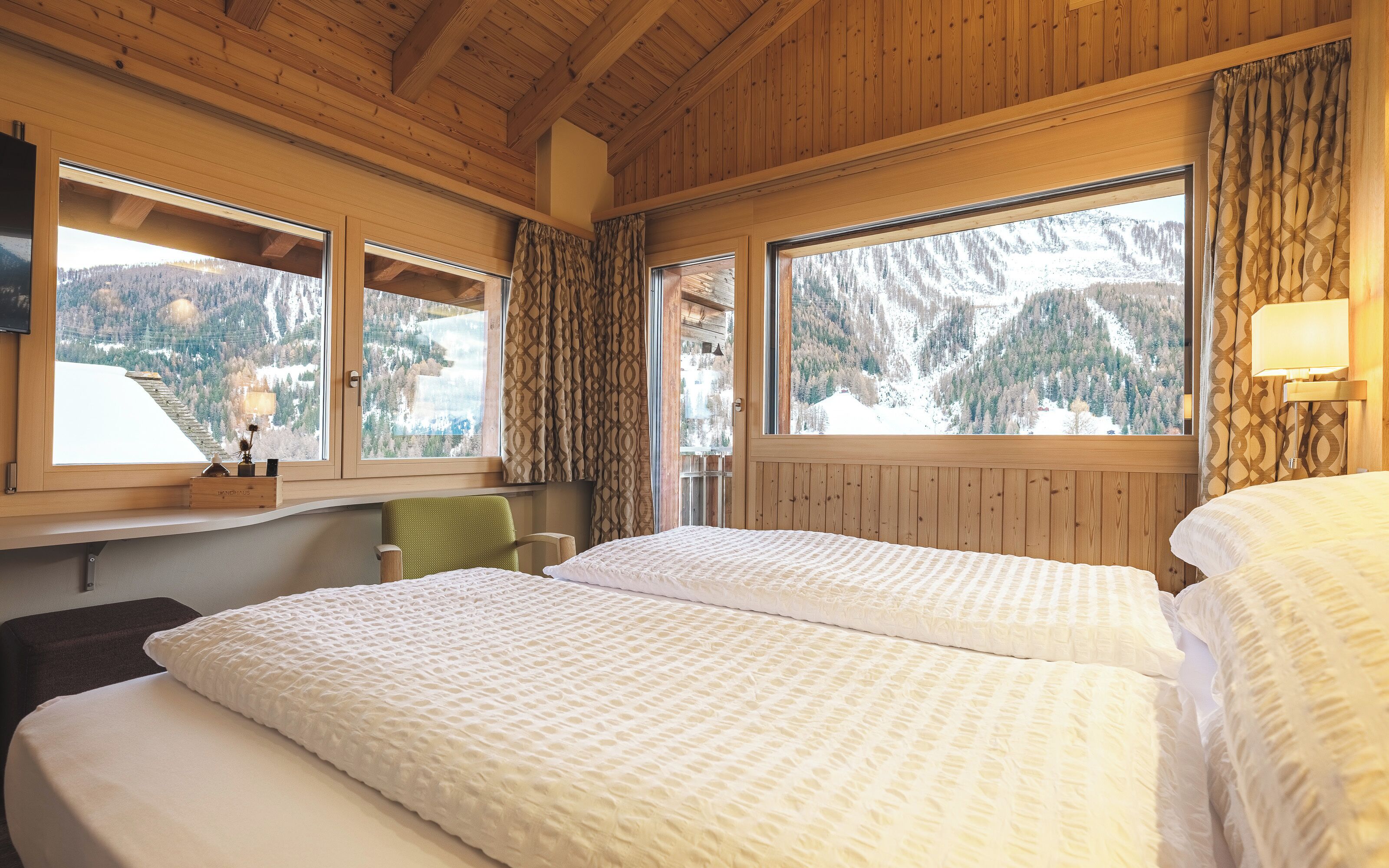 A room with two beds, wooden walls, large windows, a wooden ceiling, and a view of a snowy mountain landscape.