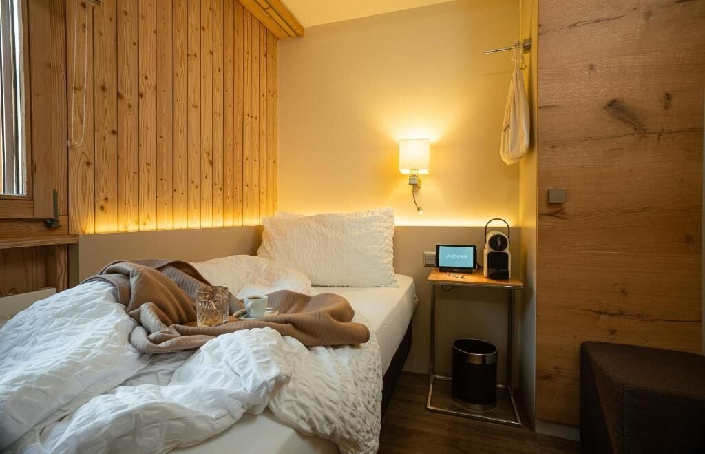 A bedroom with a bed, a bedside table with a lamp, a clock, and a small table with a cup and a glass.