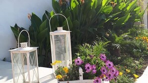 Un espace jardin agrémenté d’un bac à fleurs blanc sur lequel est inscrit le mot « MAISON », contenant des fleurs violettes et une lanterne.