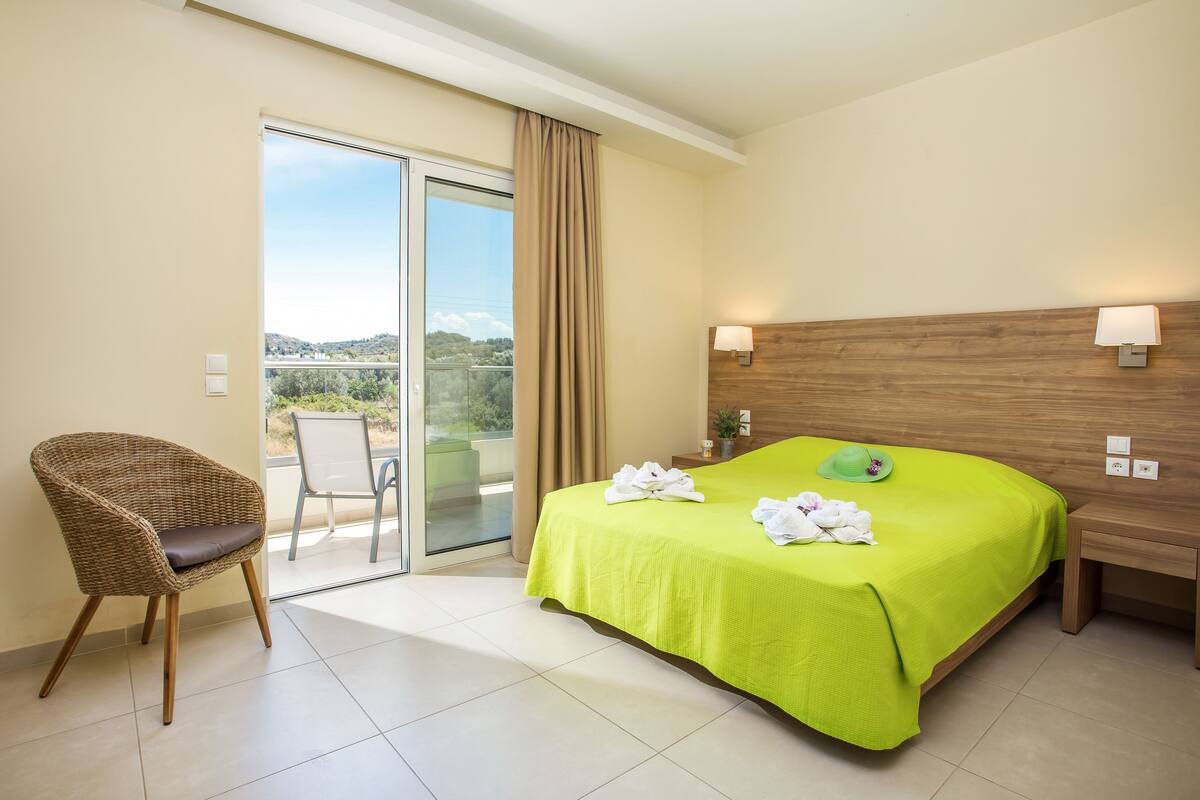 Ein Hotelzimmer mit Bett, Sessel, Nachttisch, Lampe und Blick ins Freie.