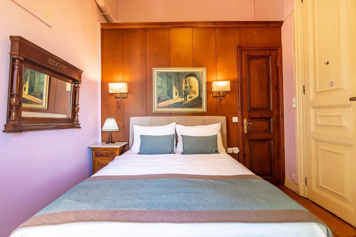 Un lit bien fait, avec deux oreillers, une tête de lit en bois et un tableau au-dessus. Il y a une table de chevet avec une lampe, et une porte sur le côté droit.