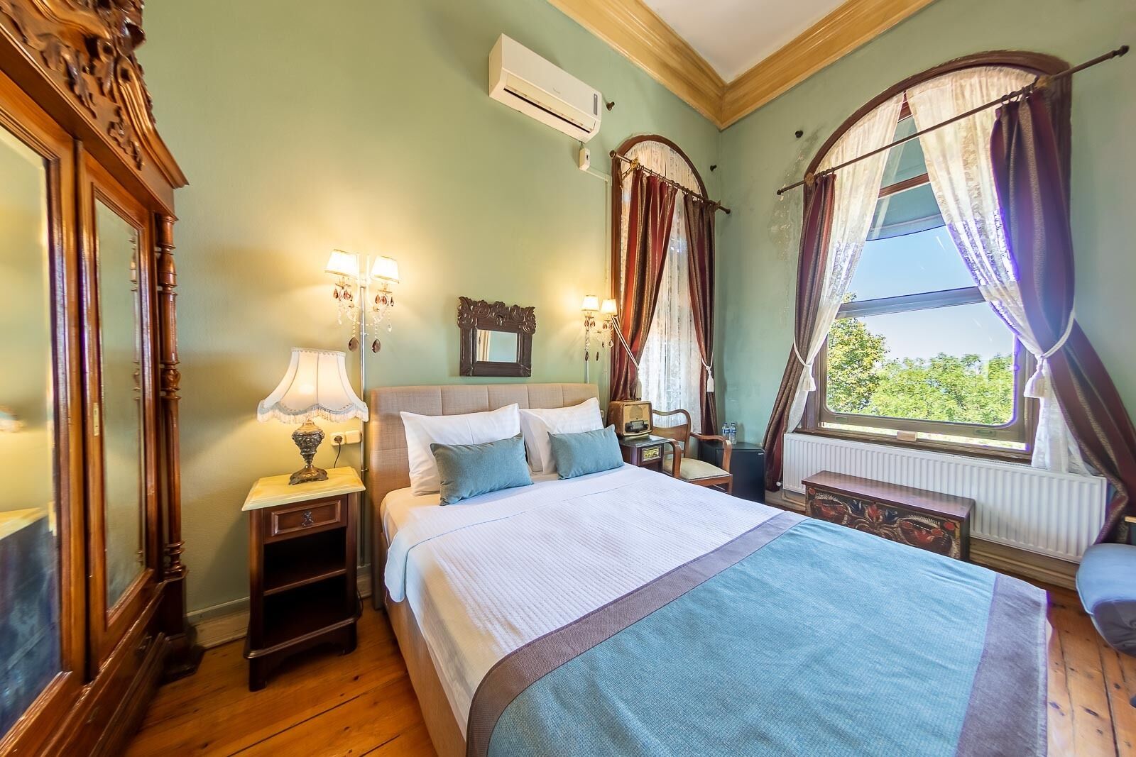 Une chambre avec un grand lit, un plancher en bois, une fenêtre avec des rideaux, un miroir et un climatiseur fixé au mur.
