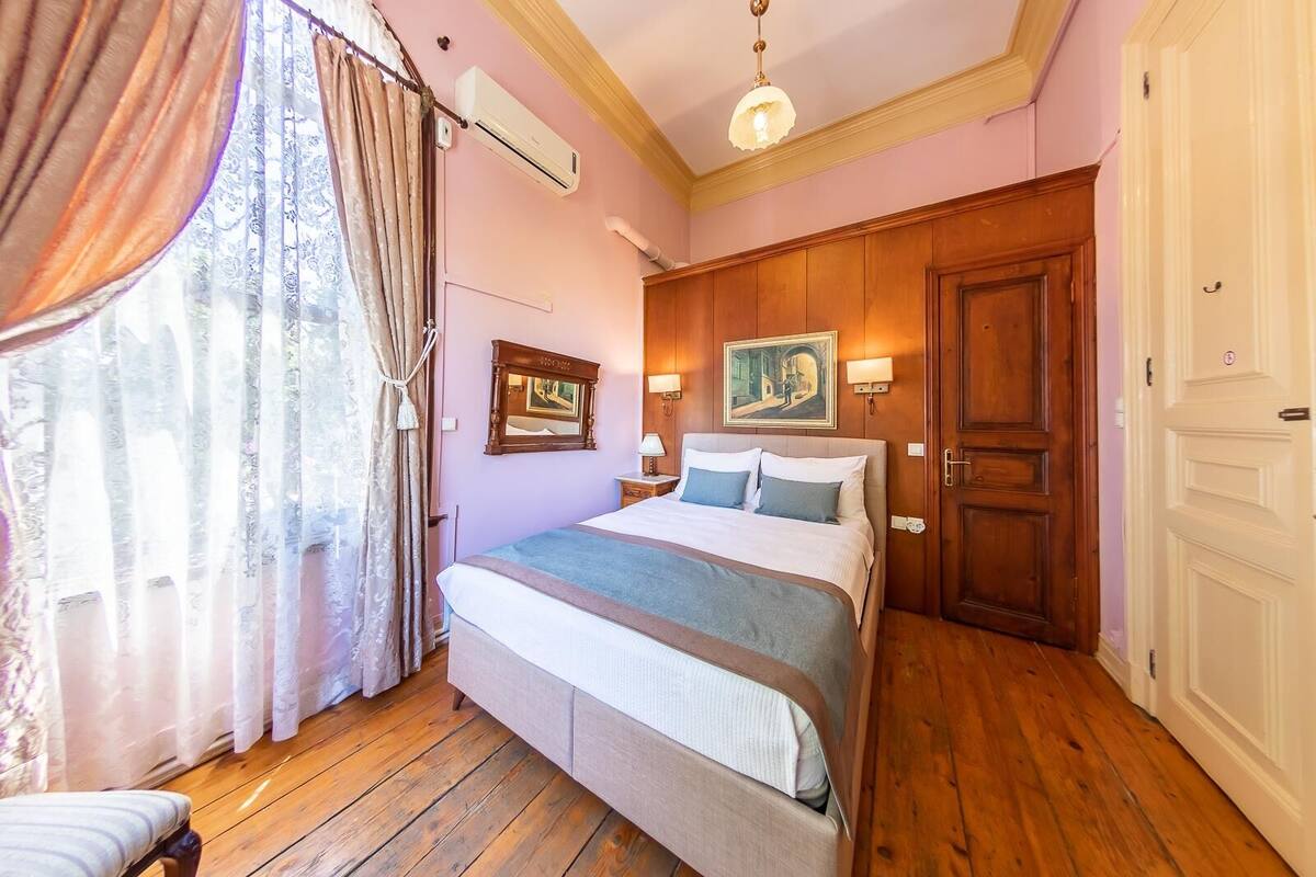 Une chambre avec un plancher en bois, un lit recouvert d’une couvre-lit bleue et blanche, une table de chevet avec une lampe, un tableau accroché au mur et une fenêtre avec des rideaux.