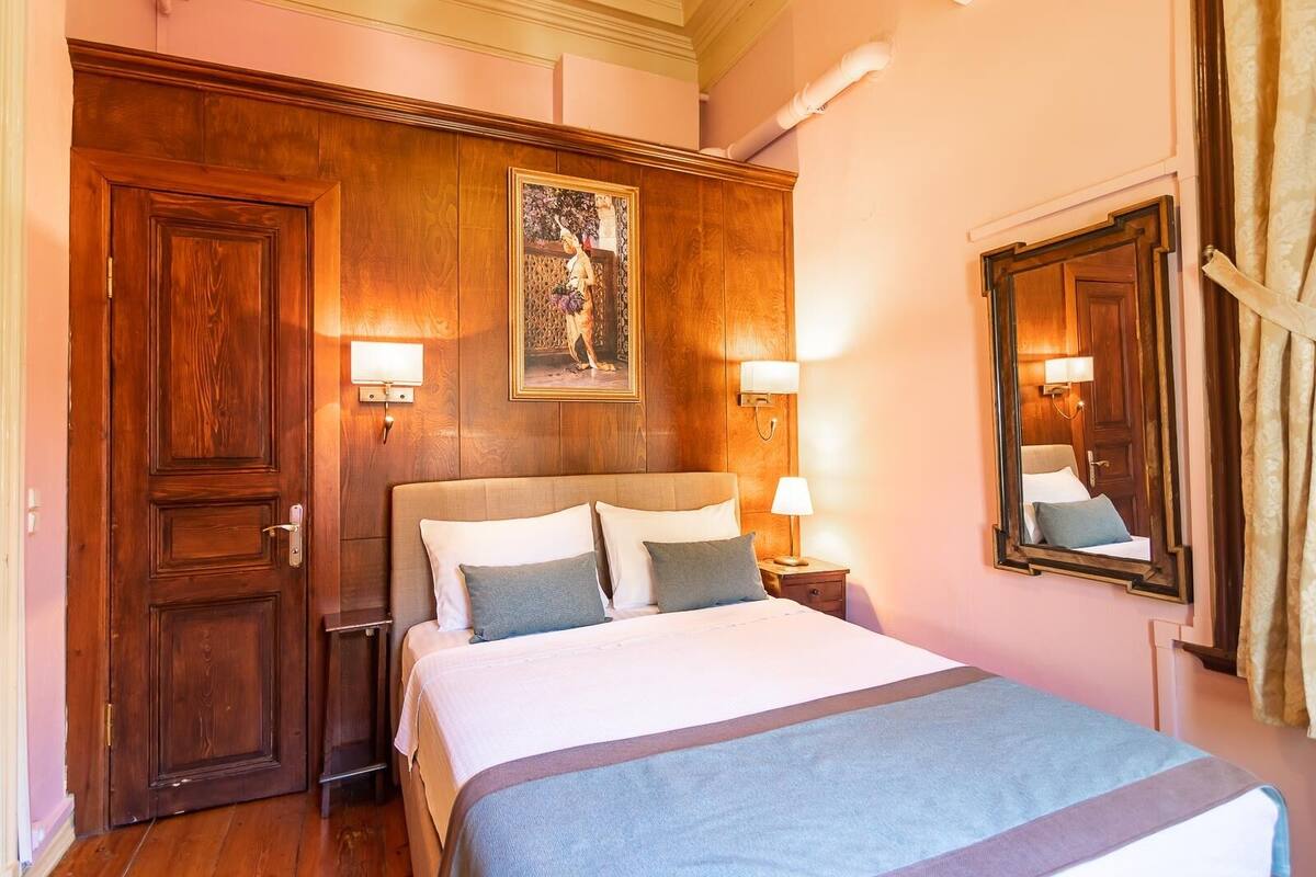 Une chambre d’hôtel avec une porte en bois, un lit avec du linge de lit blanc, deux lampes de chevet et un grand miroir au mur.