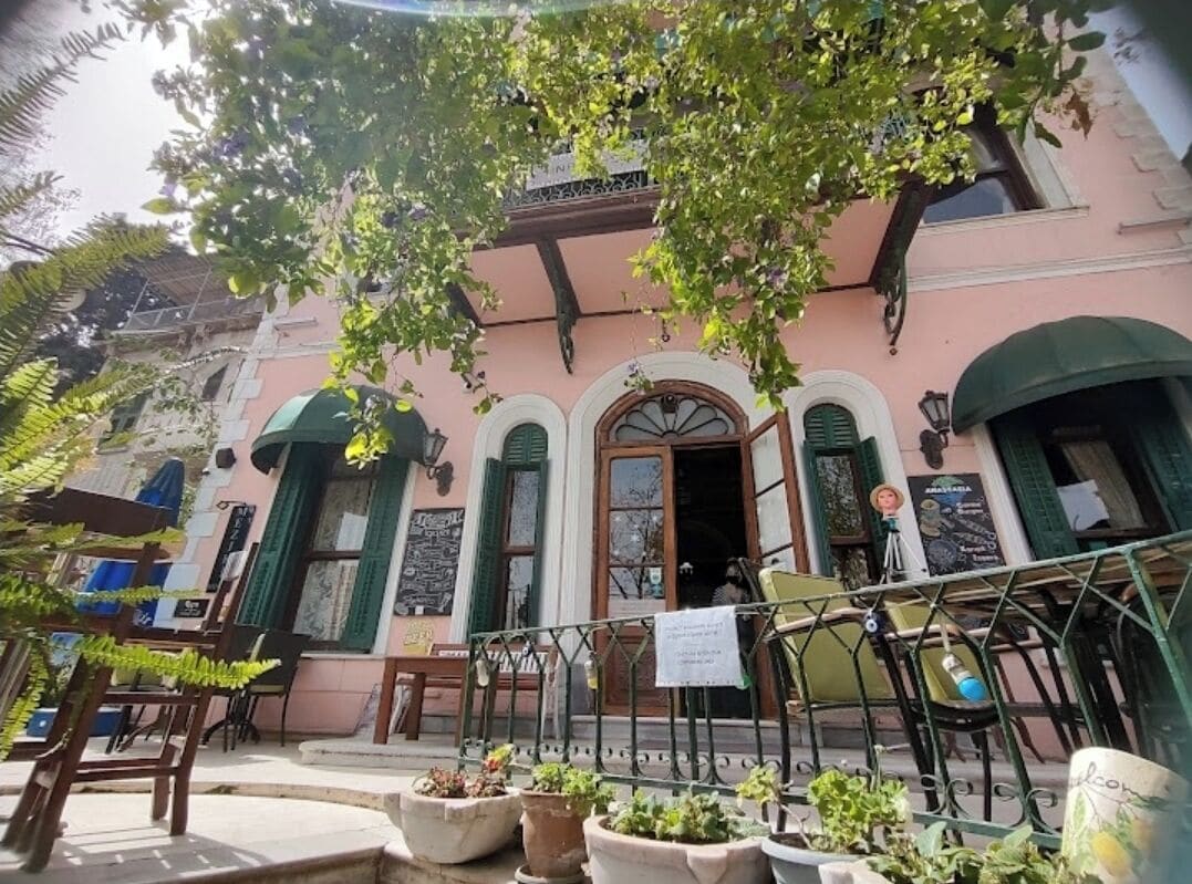 Un bâtiment à la façade rose, aux volets verts et doté d’un balcon orné de plantes en pot.