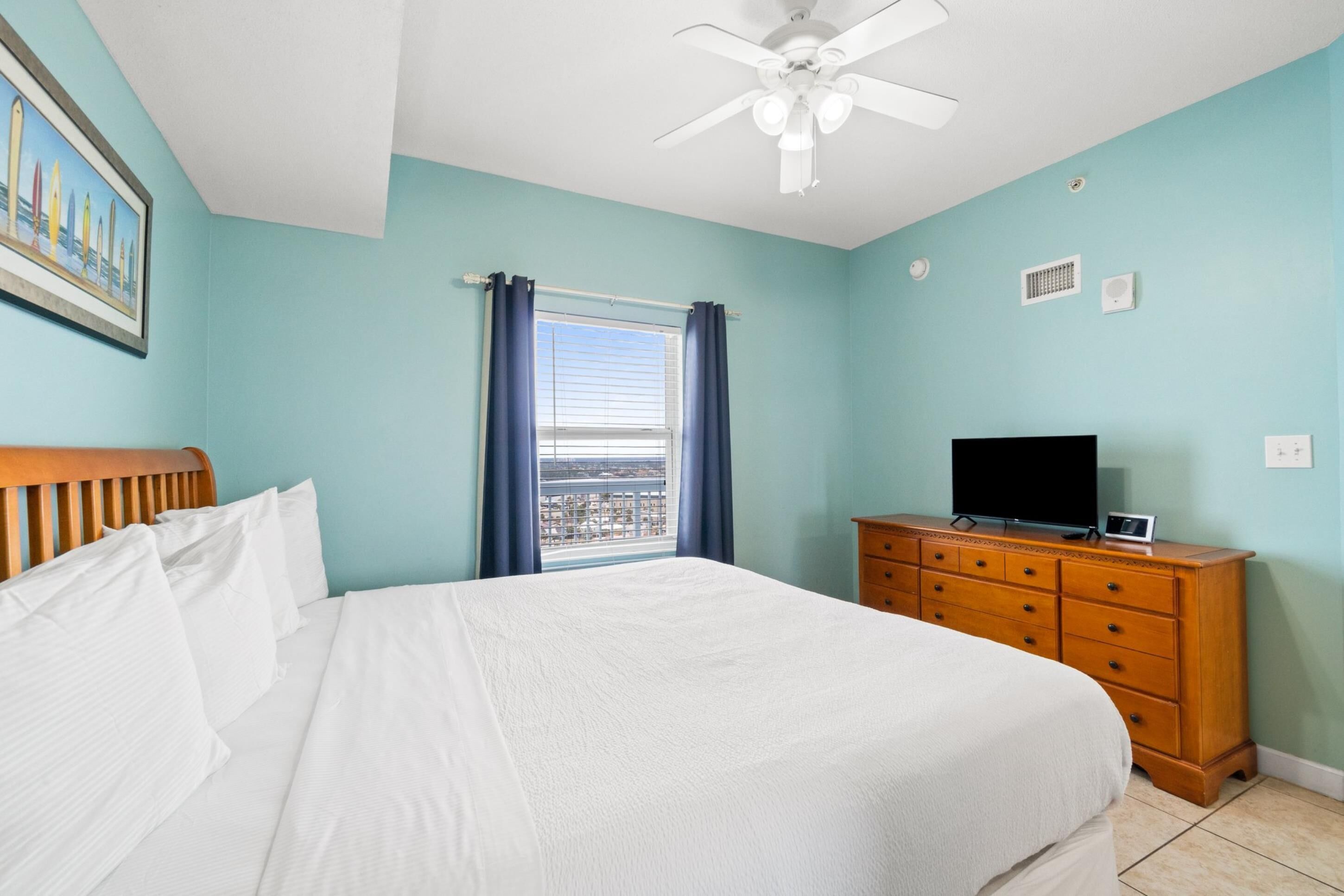 A bedroom with a bed, a dresser, a television, a ceiling fan, and a window with curtains.