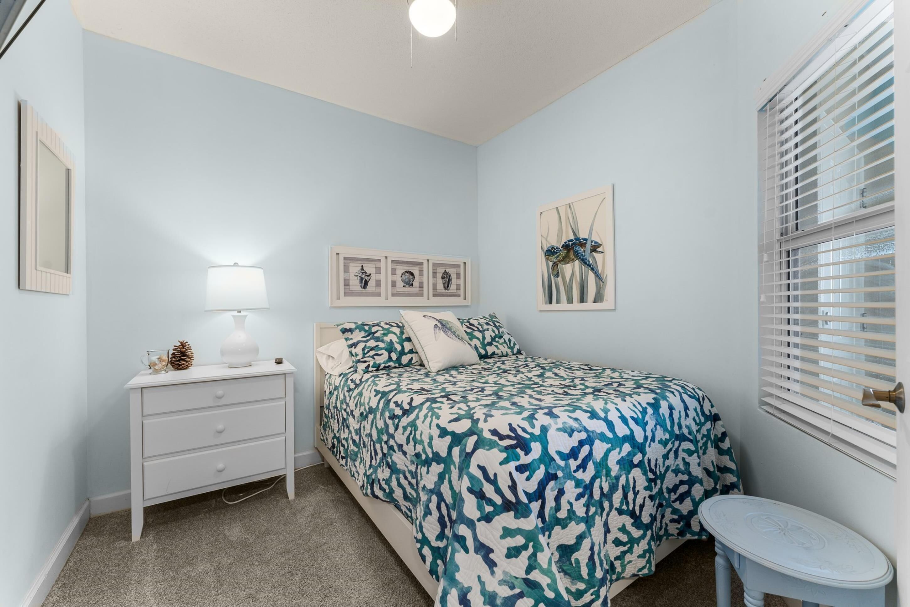 A bedroom with a bed, a nightstand, a lamp, a small table, and a window with blinds.