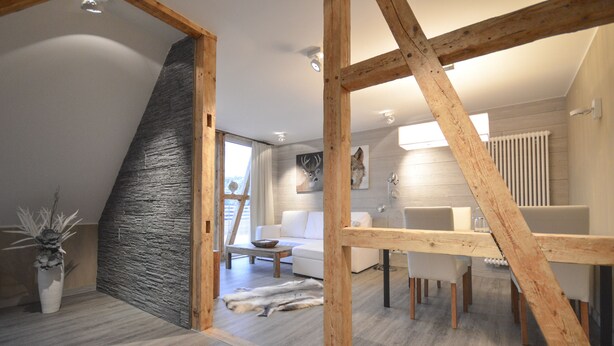 A modern loft-style living space with exposed wooden beams, a stone accent wall, a white sofa, a dining table, and a fur rug. - Stadt Chalet (Braunlage)