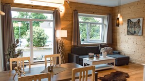 A cozy living room with a sofa, coffee table, and dining table. There are large windows with curtains, a lamp, and a painting of a wolf on the wall. - Stadt Chalet (Braunlage)