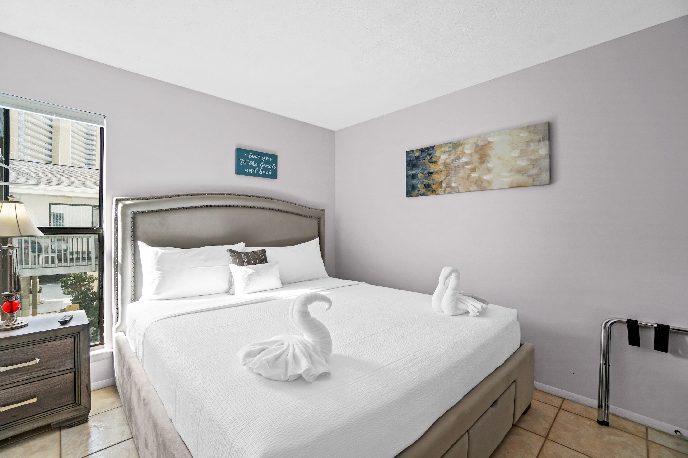 A neatly made bed with white linens and two swan-shaped towels, a bedside table with a lamp, and a view of the outside balcony.