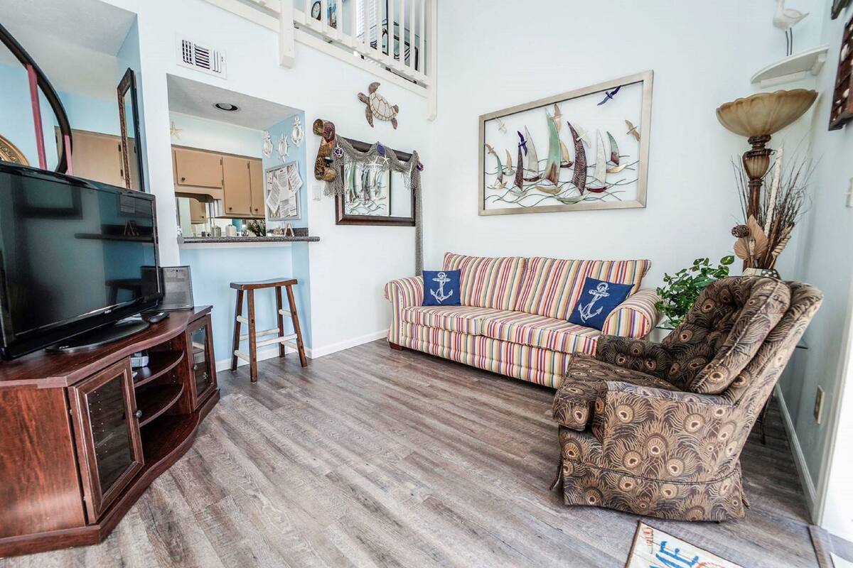 A living room with a striped sofa, a patterned armchair, a television on a wooden cabinet, and nautical-themed wall art.