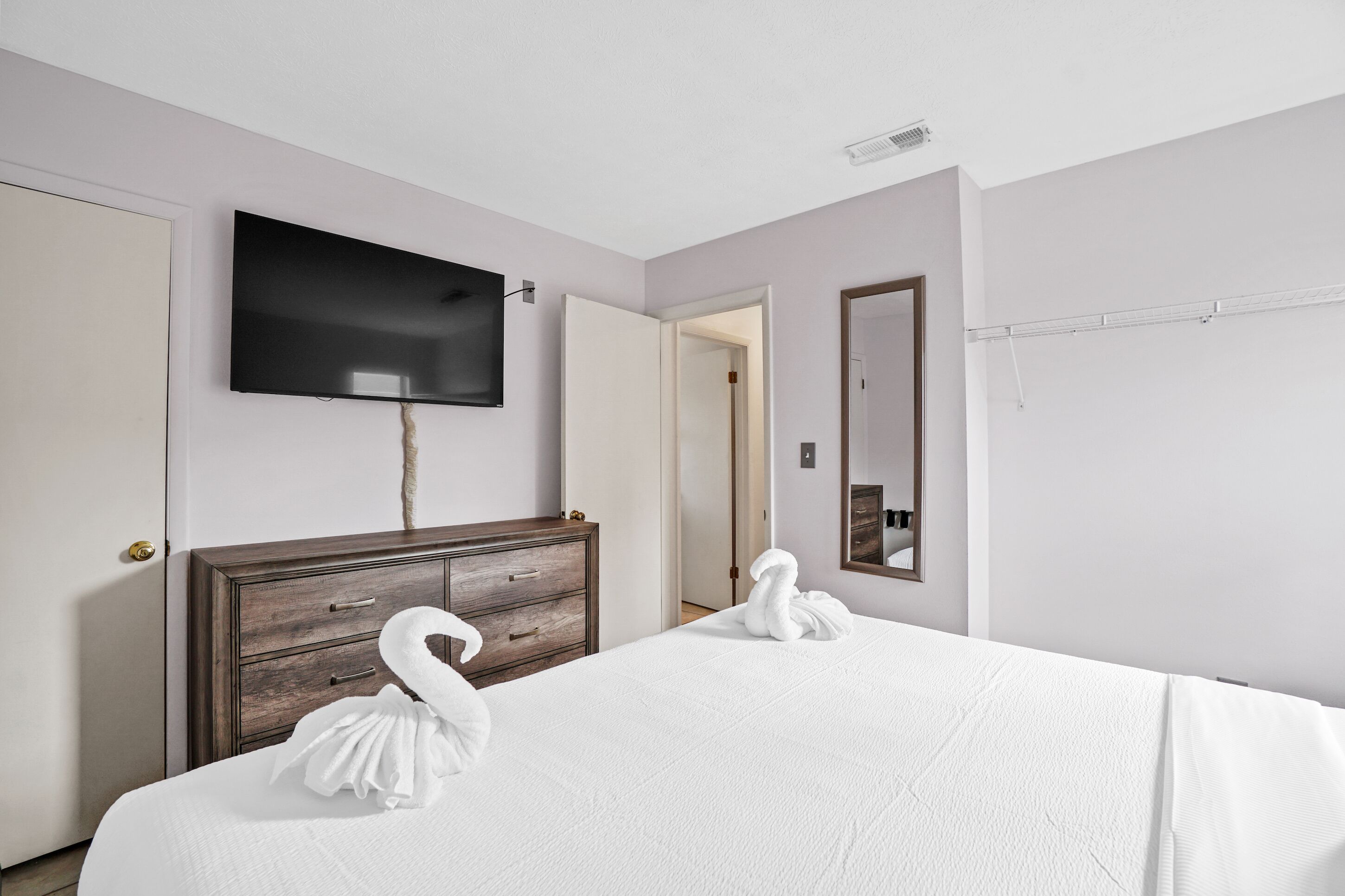 A hotel room with a bed, a wooden dresser, a flat-screen TV, a mirror, and a door.