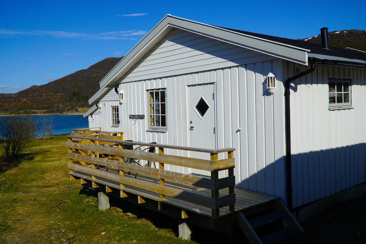 A white wooden cabin with a wooden deck and a small window, located near a body of water with mountains in the background.