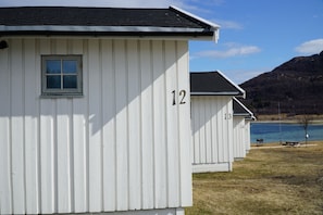 A row of white cabins numbered 12 to 15, with a lake and mountains in the background. - Tjeldsundbrua Camping (Tjeldsund)