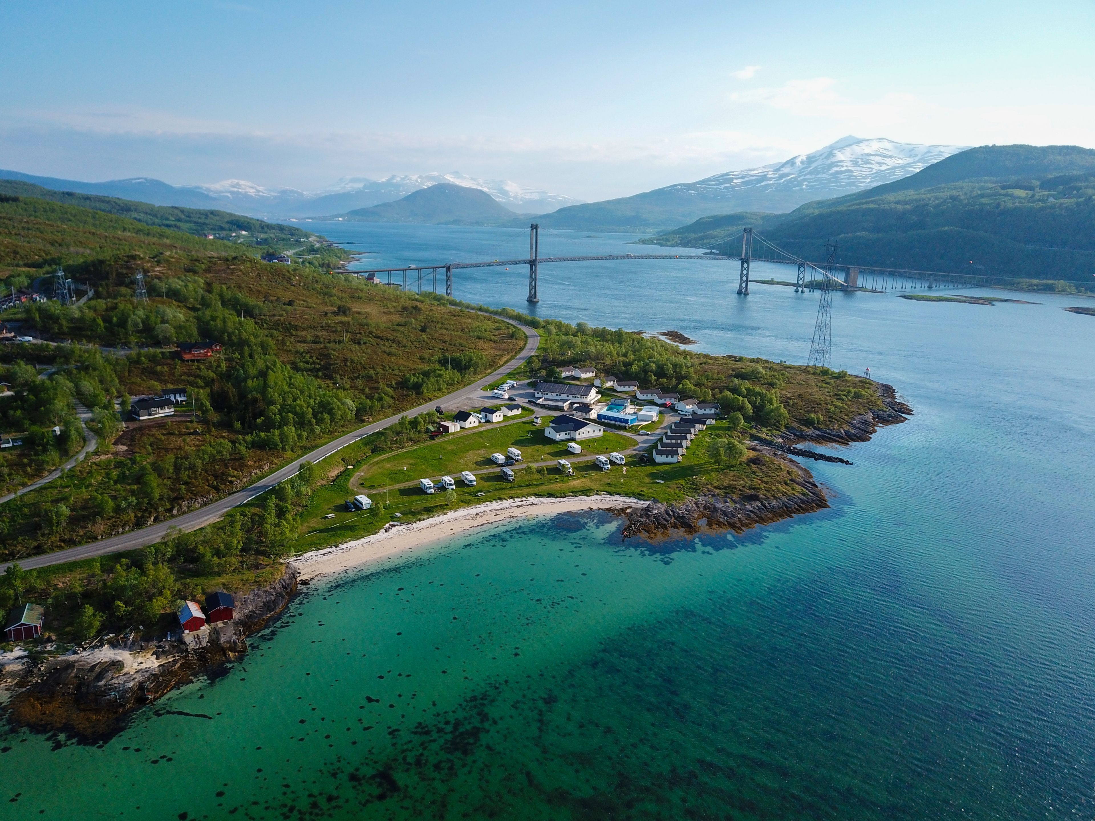 An aerial view of a coastal area with a bridge, a marina with boats, and a small settlement with houses and a road.