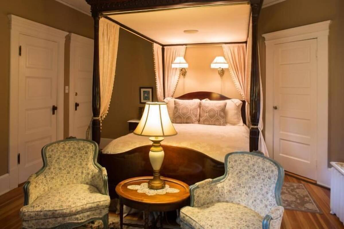 Una cama con dosel y cuatro columnas, dos lámparas de noche y sillones estampados con motivos florales.
