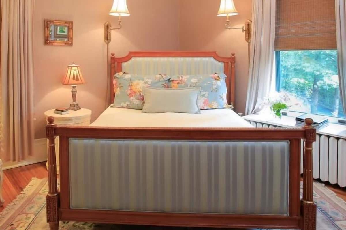 Un dormitorio con cama de madera, dos lámparas de noche, una ventana con cortinas, una mesita con lámpara y un cuadro en la pared.