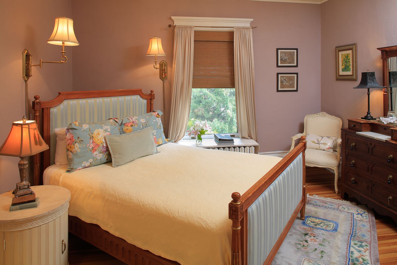 Um quarto com cama, luminárias de cabeceira, uma cadeira, cômoda e janela com cortinas.