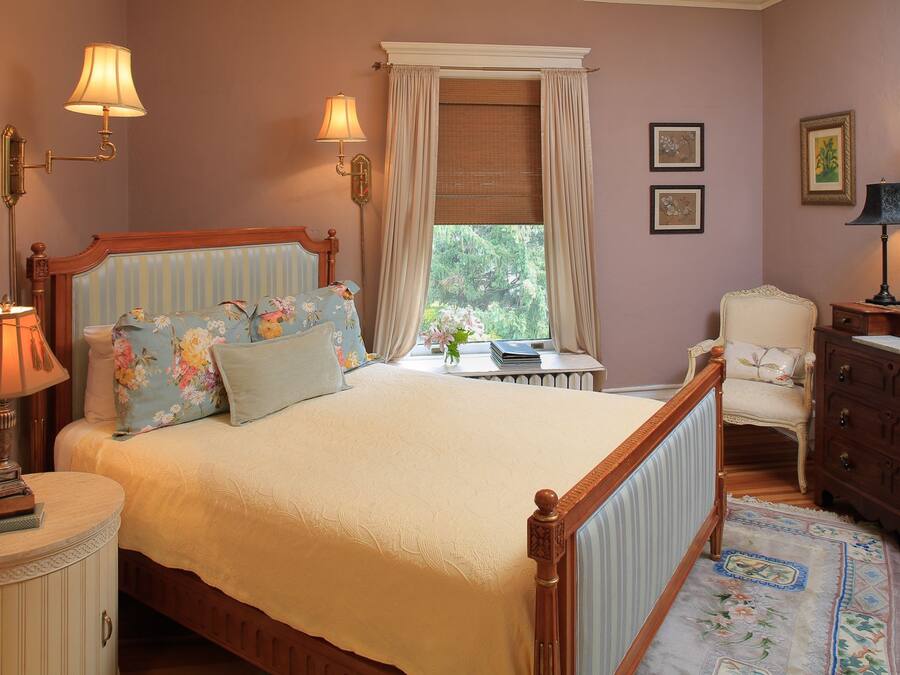 Um quarto com cama, luminárias de cabeceira, uma cadeira, cômoda e janela com cortinas.