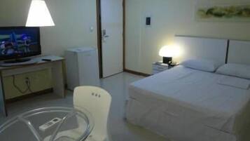 Habitación de hotel con una cama, un escritorio con televisor, una silla y una mesita de noche con lámpara.